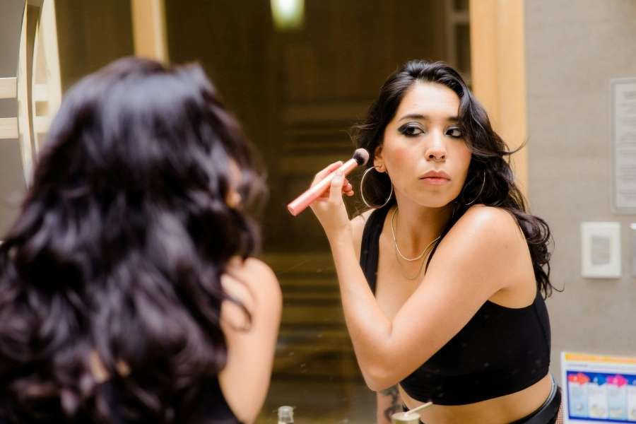 Jeune femme aux cheveux noirs appliquant un pinceau blush devant un miroir de salle de bain pour adopter un maquillage naturel au quotidien.