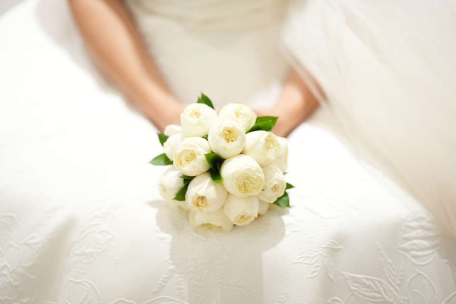 Une mariée tient entre ses mains un élégant bouquet de mariée rond composé de roses blanches serrées et de petites feuilles vertes.