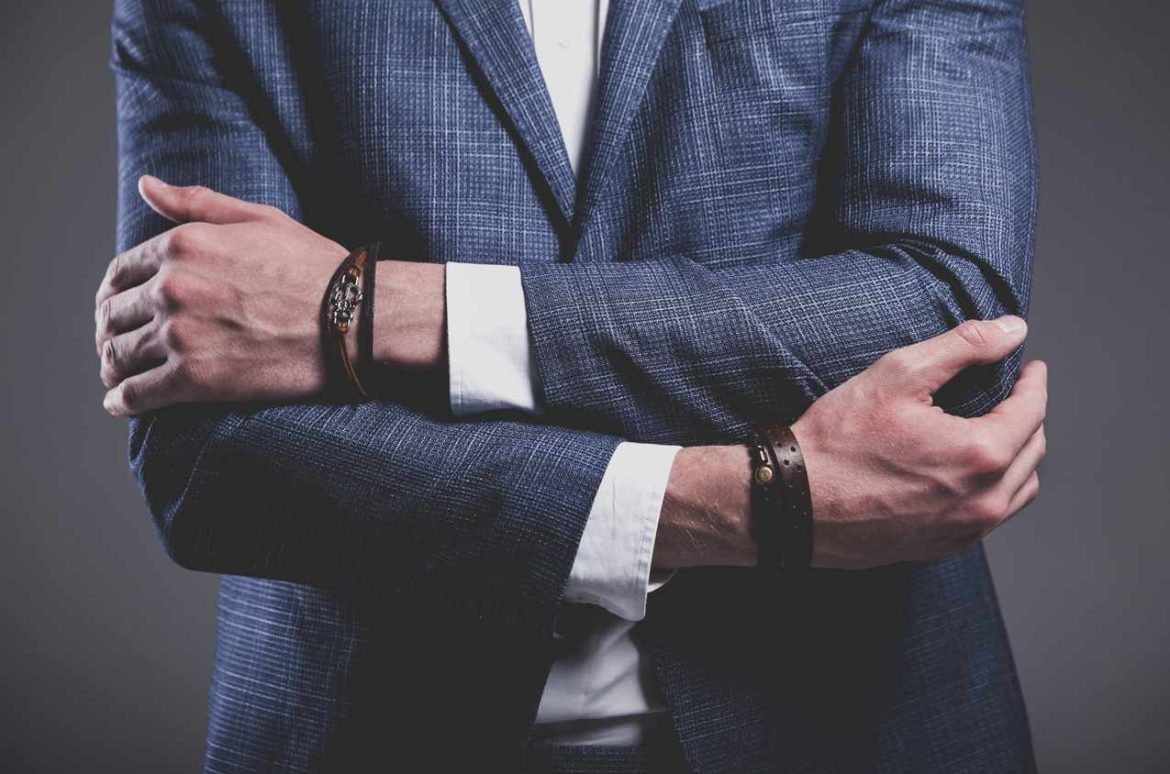 Gros plan sur les mains croisées d'un homme portant un élégant blazer bleu texturé et des bracelets en cuir, dont l'un comporte des éléments métalliques rappelant un style bracelet homme acier.
